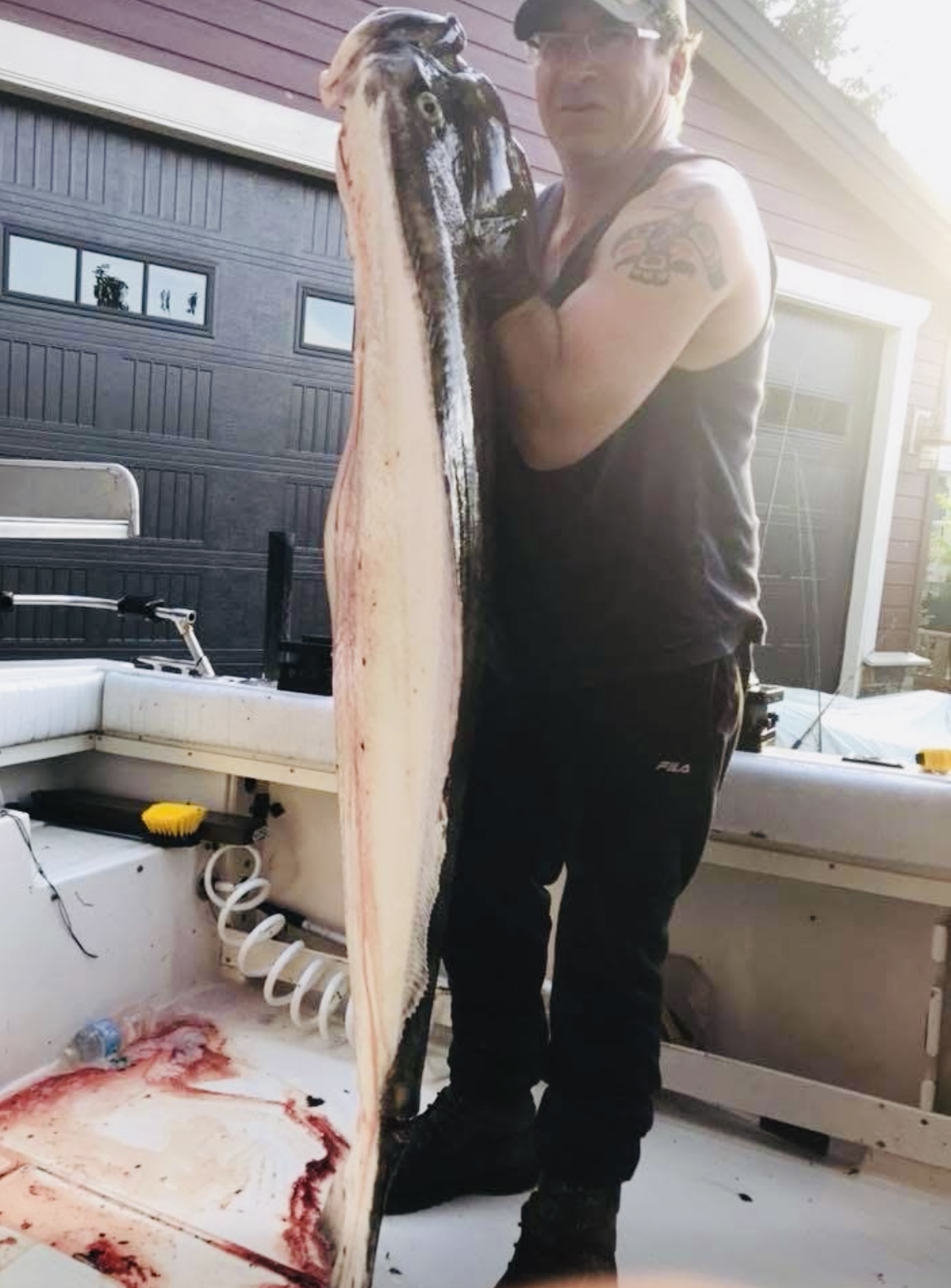 Douglas holding a large flat fish he caught, on a boat in front of a house. The fish has been cleaned and gutted, a small amount of blood is visible on the fish and the boat deck.